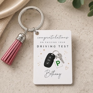 May include: A rectangular white keyring featuring a pink tassel and a silver ring. The keyring displays the text 'congratulations ON PASSING YOUR DRIVING TEST', a graphic of a car key fob with a green 'P' tag, and the name 'Bethany' in script.