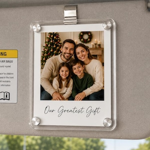 May include: A clear acrylic photo frame attached to a grey car sun visor with a metal clip. The frame holds a photograph of a family of four and features the printed text Our Greatest Gift at the bottom, secured by four silver-toned corner screws.