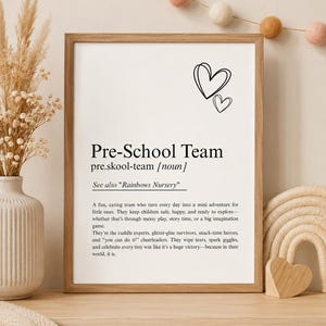 May include: A framed white print featuring black text that defines the term Pre-School Team as a noun. The text includes a description of a caring team, a reference to Rainbows Nursery, and two hand-drawn heart outlines at the top right corner.