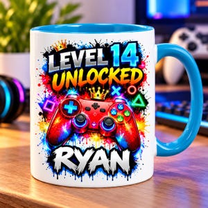 May include: A white ceramic mug with a bright blue handle and interior. The design features a red video game controller, a gold crown, and colourful paint splatters. Text reads LEVEL 14 UNLOCKED and RYAN in bold, stylised white and yellow lettering.