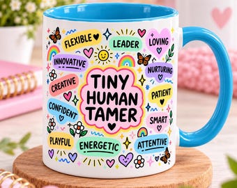May include: A white ceramic mug with a bright blue interior and handle. The exterior features colorful graphics including butterflies, rainbows, flowers, hearts, and the text "TINY HUMAN TAMER" surrounded by words like "FLEXIBLE," "LEADER," and "CREATIVE."