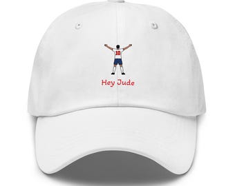 以下が含まれることがあります： 背中を向けたサッカー選手のグラフィックが描かれた白い野球帽。選手は赤い背番号10の白いジャージと青いショートパンツを着用しています。グラフィックの下には、赤いサンセリフ体で「Hey Jude」という文字がプリントされています。