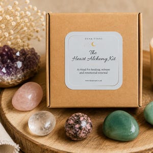May include: A square brown cardboard box labeled The Heart Alchemy Kit with a ritual for healing, release and emotional renewal. The box is surrounded by a cluster of purple amethyst, a pink rose quartz, clear quartz, rhodonite, and green stones.