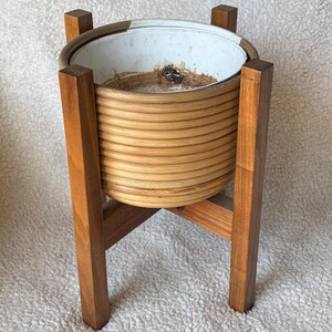 May include: A vintage plant stand with a light brown woven basket-style planter. The planter sits on a wooden stand with four legs. The stand is made of dark brown wood. The planter is empty.