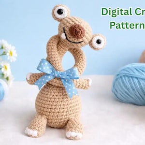 Può includere: Un bradipo giocattolo all'uncinetto color marrone chiaro con collo lungo, grandi occhi bianchi con pupille nere, naso marrone e un nastro azzurro a pois legato al collo. Il testo Digital Crochet Pattern PDF è visibile nell'angolo in alto a destra.
