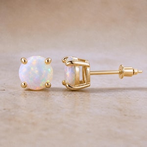 May include: A pair of stud earrings featuring round, iridescent white opals set in polished yellow gold. One earring is shown from the front, displaying the circular stone, while the other is shown from the side, revealing the gold post and backing.