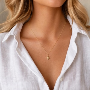 May include: A gold necklace featuring a starburst pendant. The fine chain and pendant are set against a white collared shirt. The starburst design has multiple points radiating from a central point.