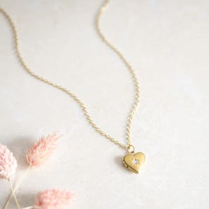 May include: A gold-toned necklace featuring a small heart-shaped locket pendant. The locket is suspended from a delicate chain and is accented with a single, tiny, clear, round-cut stone set in the centre, surrounded by a subtle starburst engraving.