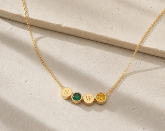 May include: A gold-toned chain necklace featuring four small circular charms. Two charms are engraved with the letters S and W, while the other two charms are set with a round green gemstone and a round yellow gemstone respectively.