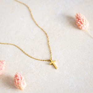 May include: A gold-coloured necklace featuring a simple, plain cross pendant suspended from a delicate cable chain. The metal has a smooth, polished finish, and the cross is centred on the chain, which extends upwards and out of the frame.