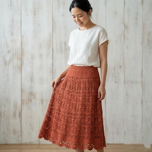 May include: A short-sleeved white top paired with a rust-coloured midi skirt featuring an intricate, openwork crochet pattern. The skirt has a fitted waistband and a scalloped hemline, showcasing a detailed, repeating floral-inspired knit design.