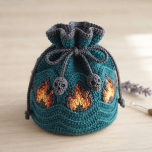 Puede incluir: Una bolsa de ganchillo con cordón en color verde azulado que presenta un patrón repetitivo de llamas en naranja y amarillo. La bolsa se cierra con un cordón gris oscuro que termina en dos pequeñas cuentas tejidas con forma de calavera.