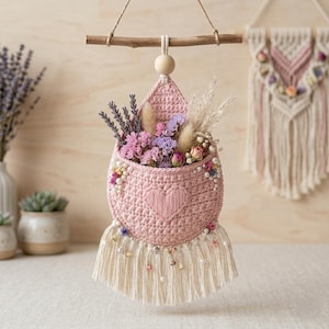 May include: A pink crocheted wall hanging pocket featuring a heart motif, adorned with white pearls and a cream-colored fringe. The pocket is suspended from a wooden dowel and contains an arrangement of dried lavender, pink flowers, and grasses.
