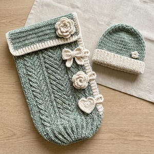 May include: A sage green crochet baby sleeping bag and matching beanie hat set. The sleeping bag features a herringbone stitch pattern, a cream-colored fold-over flap, and decorative cream crochet accents including two flowers, a bow, and a heart.