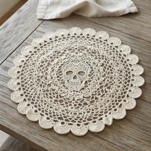 May include: A round, off-white crochet doily featuring an intricate, openwork lace pattern with a central skull motif. The circular piece has a scalloped edge with repeating fan-like details, crafted from fine thread in a delicate, decorative design.