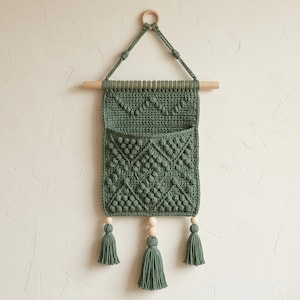 Pode incluir: Um pendurador de parede em macramé verde sálvia com um bolso e três borlas. A peça artesanal apresenta um pino e um anel de madeira para pendurar. O bolso tem um padrão de diamante texturizado. As borlas são acentuadas com contas de madeira.