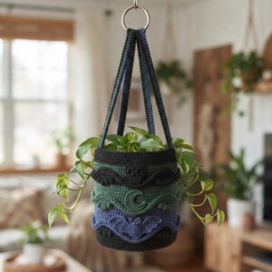 Puede incluir: Un soporte para plantas de ganchillo colgante con diseño cilíndrico y bandas horizontales en negro, verde oscuro y azul oscuro. El exterior está decorado con motivos de ganchillo en relieve que incluyen una calavera, una luna y un murciélago.