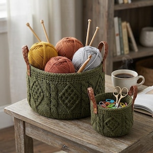 May include: Two round, olive green crocheted baskets with braided brown faux leather handles. The larger basket holds four yarn balls in mustard, rust, and gray, with wooden knitting needles. The smaller basket contains gold scissors and colorful stitch markers.