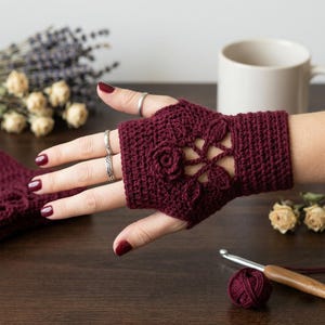 May include: A deep burgundy fingerless glove featuring a detailed crochet rose and leaf motif on the back of the hand. The glove has a ribbed cuff and an openwork design, crafted from yarn with a visible texture and consistent stitch pattern.