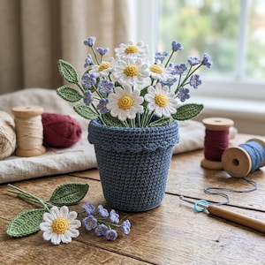 May include: A crocheted blue pot containing a bouquet of white daisies with yellow centers and small purple flowers with green leaves. Additional loose crocheted flowers and leaves lie nearby on a wooden surface with yarn spools.