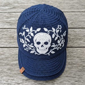 Puede incluir: Una gorra de béisbol tejida a ganchillo en color azul oscuro con un diseño de calavera blanca bordado en el centro del panel frontal, rodeado de patrones de enredaderas con hojas blancas. Una pequeña etiqueta rectangular de cuero marrón está en la visera.