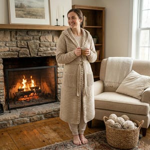 May include: A long, beige, hooded cardigan made of thick, textured knit fabric. The garment features long sleeves, two large front patch pockets, and a matching tie belt at the waist. The edges are finished with a decorative, small-beaded trim.