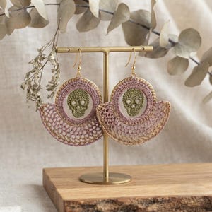 May include: A pair of dangle earrings featuring a circular crochet design in dusty pink and gold thread. Each earring centers an olive green crocheted skull motif within the circle, suspended from gold-toned metal hooks on a gold display stand.