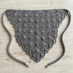Puede incluir: Un pañuelo triangular gris tejido a ganchillo con un borde festoneado. El pañuelo está adornado con pequeñas cuentas plateadas y botones decorativos. Tiene largas cintas de ganchillo a cada lado para atar al cuello.