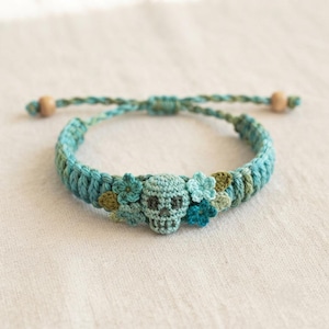 May include: A teal crocheted bracelet featuring a central skull motif flanked by small floral accents in shades of blue and green. The band is constructed from braided cord with two light brown wooden beads at the ends of the adjustable ties.