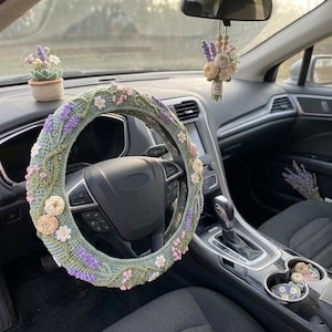 May include: A sage green crochet steering wheel cover decorated with embroidered lavender sprigs, small white daisies, and pale pink flowers. The set includes a matching crochet potted plant, a hanging floral car charm, and small floral cup holder inserts.