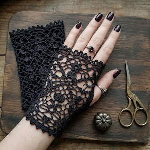 May include: A pair of black fingerless gloves featuring an intricate crochet lace pattern with floral motifs and scalloped edges. One glove is worn on the hand, while the other lies flat, accompanied by a small metal pumpkin and stork scissors.