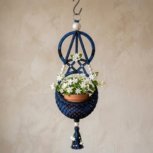 May include: A hanging macrame plant holder in dark blue and black cord, featuring a circular top frame, white pearl-like beads, and a bottom tassel. It holds a terracotta pot containing a small plant with white star-shaped flowers and green leaves.