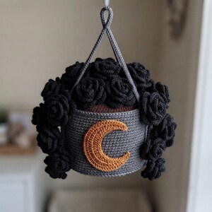 May include: A hanging crochet planter featuring a dark gray cylindrical pot with a prominent golden-brown crescent moon appliqué on the front. The planter is filled with numerous black crocheted roses and suspended by a matching gray cord loop.