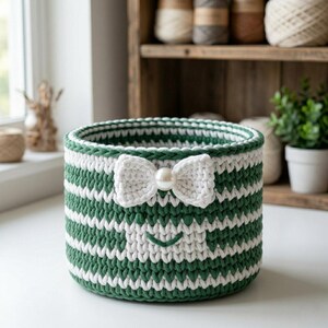 May include: A round, crocheted storage basket featuring horizontal stripes in dark green and white. The front of the basket is decorated with a white crocheted bow centered with a single white pearl bead and a small, stitched green smile below.