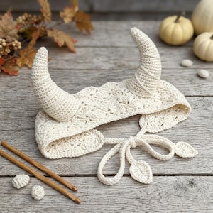 May include: An off-white crochet headband featuring two curved, pointed horns extending upward. The headband is crafted with a scalloped stitch pattern and includes long, thin ties at the back that end in small, leaf-shaped accents for secure fastening.
