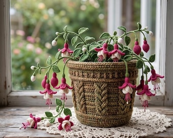 Könnte beinhalten: Eine gehäkelte Fuchsie mit leuchtend pinken und weißen Blüten sowie grünen Blättern in einem strukturierten, braunen Häkeltopf. Die Pflanze zeigt zahlreiche hängende Blüten und Knospen, die aufwendige Handarbeit und detaillierte Maschenmuster aufweisen.