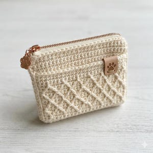May include: A cream-colored crocheted pouch featuring a diamond-patterned texture on the front pocket. The pouch includes a rose gold zipper with a paw print charm and a small tan leather tab embossed with a paw print on the front pocket edge.