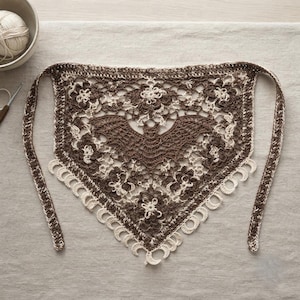 May include: A triangular crochet bandana featuring a central dark brown bat motif surrounded by intricate floral lace patterns in cream and brown yarn. The bottom edge is finished with a series of small, cream-colored crescent moon shapes.