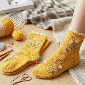 Può includere: Un paio di calzini in maglia color giallo senape con motivi floreali ricamati dai petali bianchi, blu e viola. I calzini presentano un bordo smerlato color crema sul polsino. Un calzino è indossato su un piede, l'altro è appoggiato.