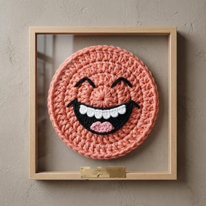 Pode incluir: Uma tapeçaria circular de crochê na cor pêssego com um rosto risonho, apresentando olhos de fio preto, uma boca larga preta, dentes brancos e uma pequena língua rosa, tudo dentro de uma moldura quadrada de madeira clara com frente de vidro.