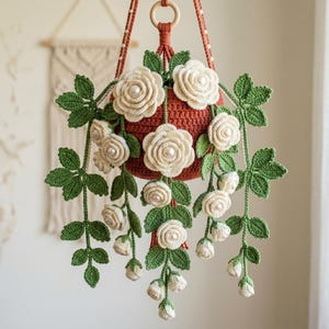 May include: A hanging crochet plant featuring a terracotta-colored pot, vibrant green crocheted leaves, and multiple cream-colored crocheted roses. Each rose is accented with a central white pearl bead. The plant is suspended by a matching cord.