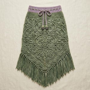 May include: A sage green crochet skirt featuring an intricate floral pattern, a lavender waistband with a matching drawstring, and a long fringe hem. The garment is crafted with detailed needlework, showcasing a textured design and a tie closure.