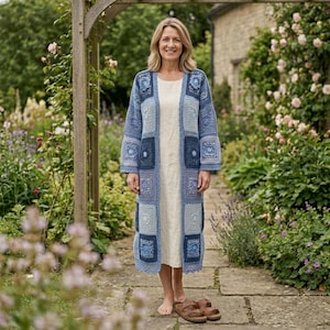 May include: A long, open-front cardigan made of various blue and white crocheted square motifs, featuring a mix of light, medium, and dark blue shades. The garment has long sleeves and a straight hem, worn over a simple, solid cream-colored dress.