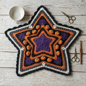 Puede incluir: Un tapete de ganchillo en forma de estrella con un centro morado, capas concéntricas naranjas y negras, y pequeñas calabazas naranjas tridimensionales. El borde incluye motivos de fantasmas blancos, rodeado por un cuenco, tijeras y un ganchillo de madera.
