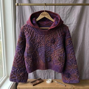 Puede incluir: Un suéter de ganchillo con capucha y mangas largas que presenta un patrón de telaraña texturizado en tonos variados de morado y marrón. La prenda incluye motivos de arañas tridimensionales integrados, un dobladillo acanalado y puños acanalados.