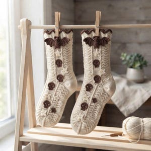 May include: A pair of cream-colored crocheted socks featuring a textured chevron pattern, adorned with small brown crocheted skulls, cream-colored stars, and a prominent brown crocheted bat motif at the top cuff, hanging from wooden clothespins.