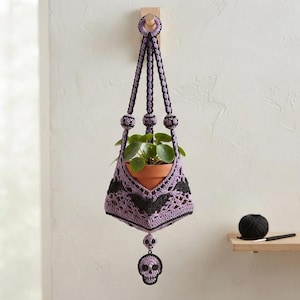 Peut inclure: Une suspension pour plante en crochet lavande et noir avec un motif de chauves-souris et une breloque crâne suspendue. Le support contient un pot en terre cuite avec une plante verte et est suspendu par une boucle en crochet assortie.