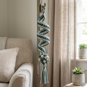 May include: A spiral-shaped crochet wall hanging featuring alternating bands of cream, sage green, and dark teal yarn. The design includes a cluster of small crocheted spheres at the base, finished with a long, multi-coloured yarn tassel.