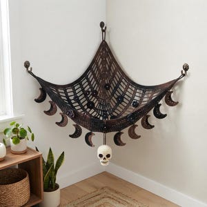 May include: A triangular crochet corner hammock in dark brown and black yarn, featuring a lattice pattern, small black floral accents, and a series of crescent moon motifs hanging along the bottom edge, with a single cream-coloured skull charm.
