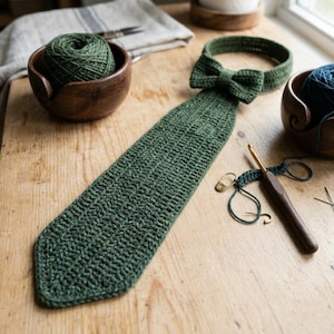 May include: A dark green crocheted necktie featuring a matching bow tie at the collar. The tie is constructed with a textured stitch pattern and is displayed alongside a wooden crochet hook, a gold stitch marker, and a ball of green yarn.